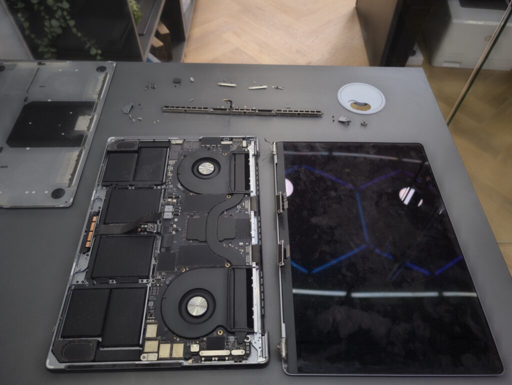 MacBook Pro 16 fully separated on the bench - main body on the left with logic board and fans, old damaged display assembly upright on the right, hinge bar and torx screws laid out above