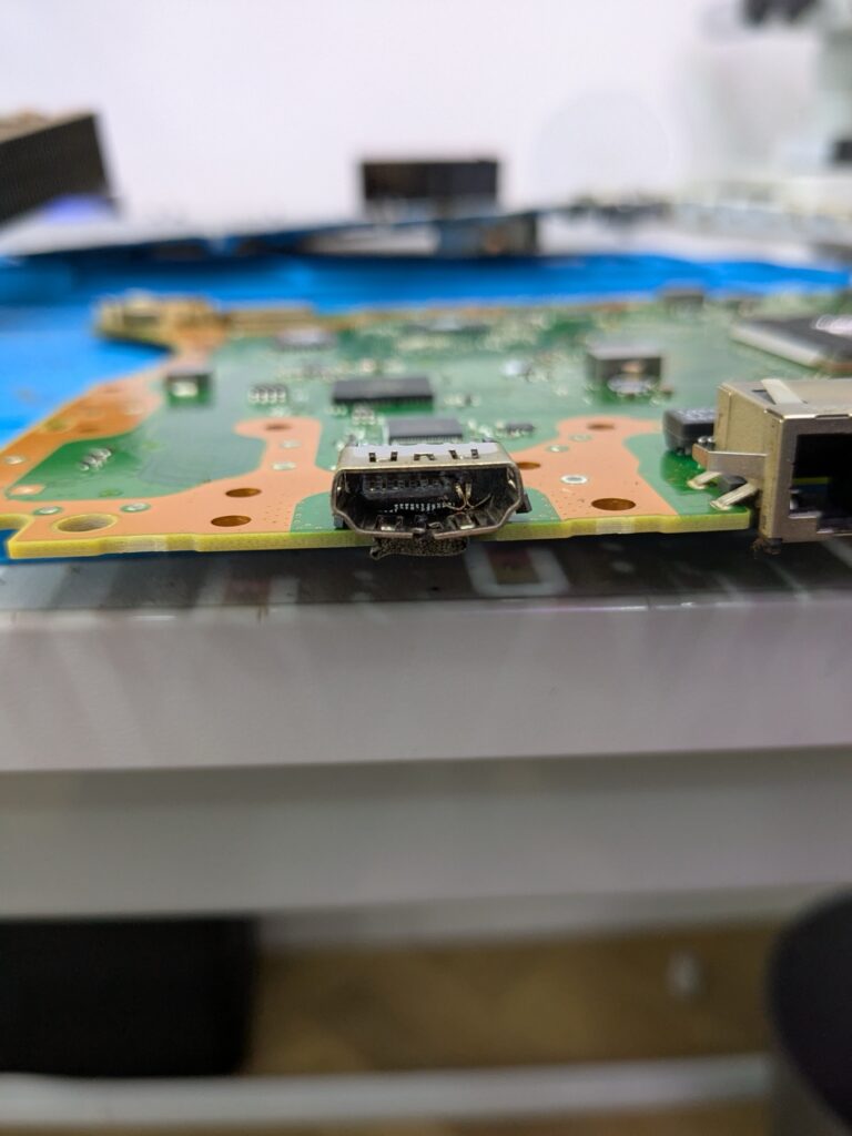 Closeup at low angle of the damaged HDMI port on the PlayStation 5 Slim logic board, showing the pins inside the socket bent flat against the back wall of the connector