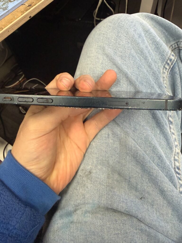 Side view of the iPhone 14 Plus showing the mute switch, volume buttons and SIM tray, with splinters of broken back glass along the edge where the back meets the frame