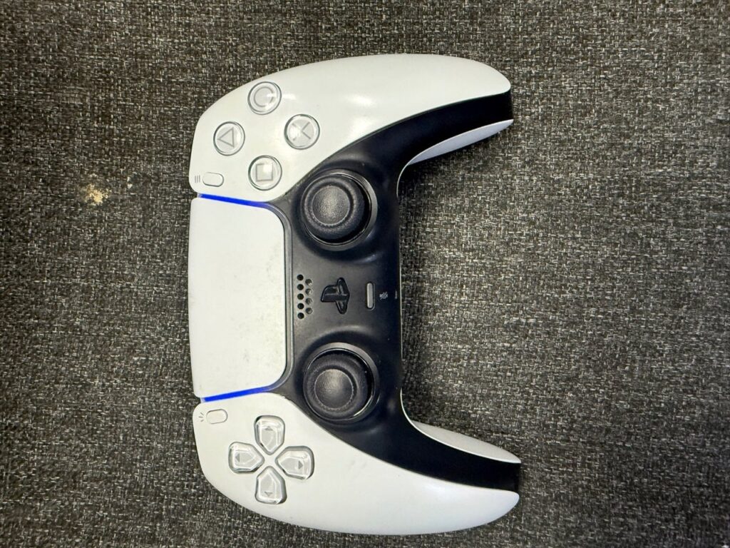 PS5 DualSense controller photographed sideways on grey fabric, showing both thumbsticks, the touchpad, the face buttons and the d-pad, with the light bar glowing blue