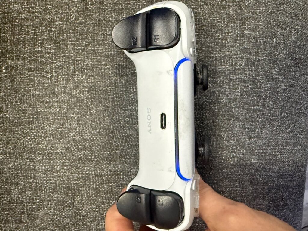 Top edge of a PS5 DualSense controller held in the hand, showing the R1, R2, L1 and L2 buttons, the USB-C charging port and the blue light bar